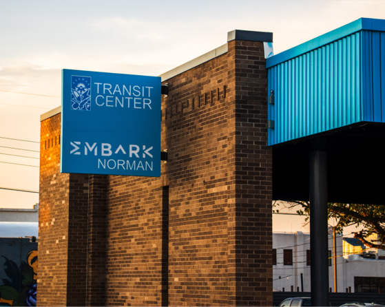 A view of the Noman Transit Center from the east showing a blue marquee sign with the City of Norman Logo the text "Transit Center" and the EMBARK  Norman logo