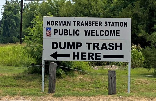 White sign with City Logo and Black Letters that say Norman Transfer Station. Public welcome. Dump trash here.